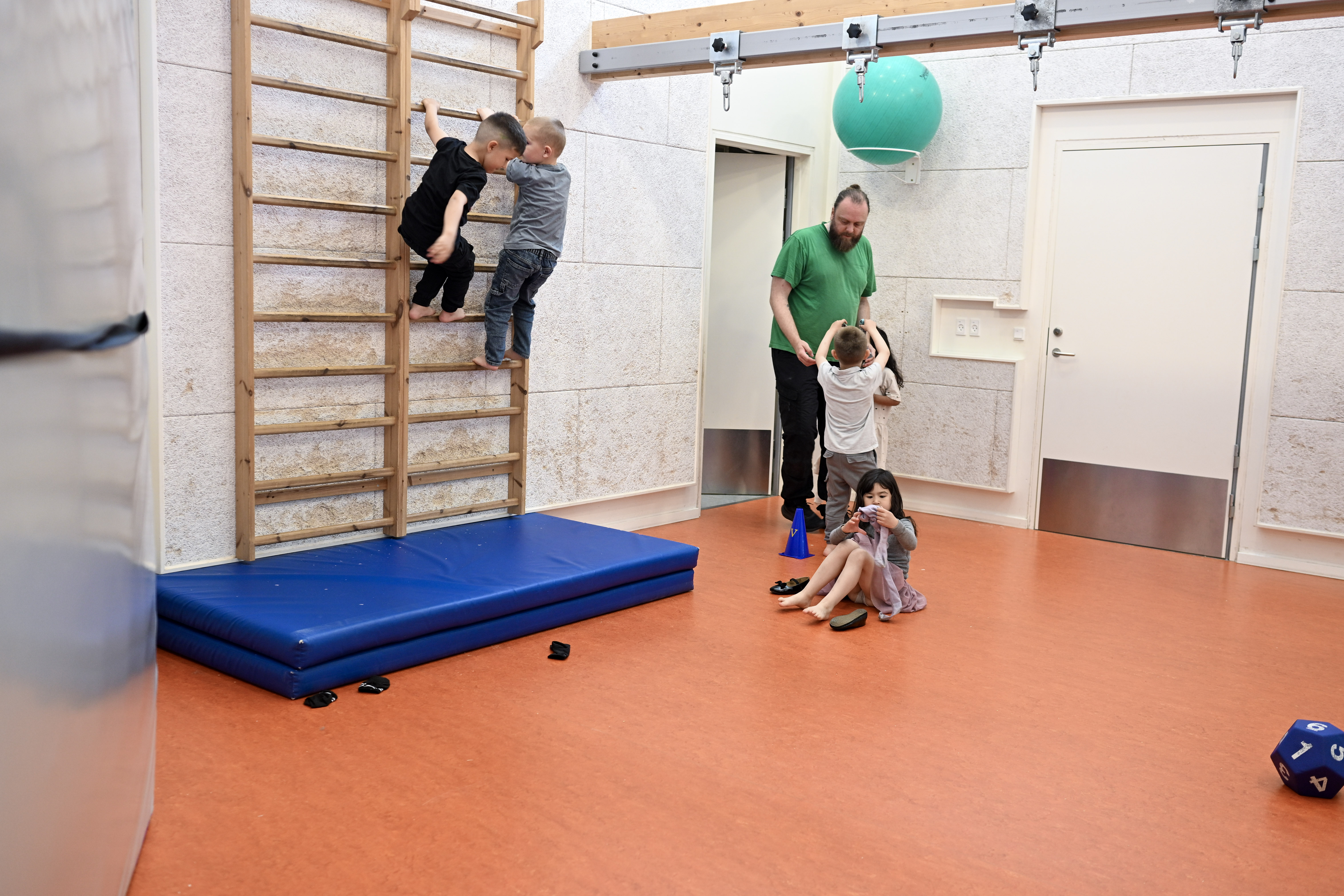 Børn klatrer i ribber og taler med voksen i gymnastiksal